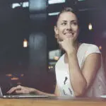 A laughing woman at a table with her open MacBook.